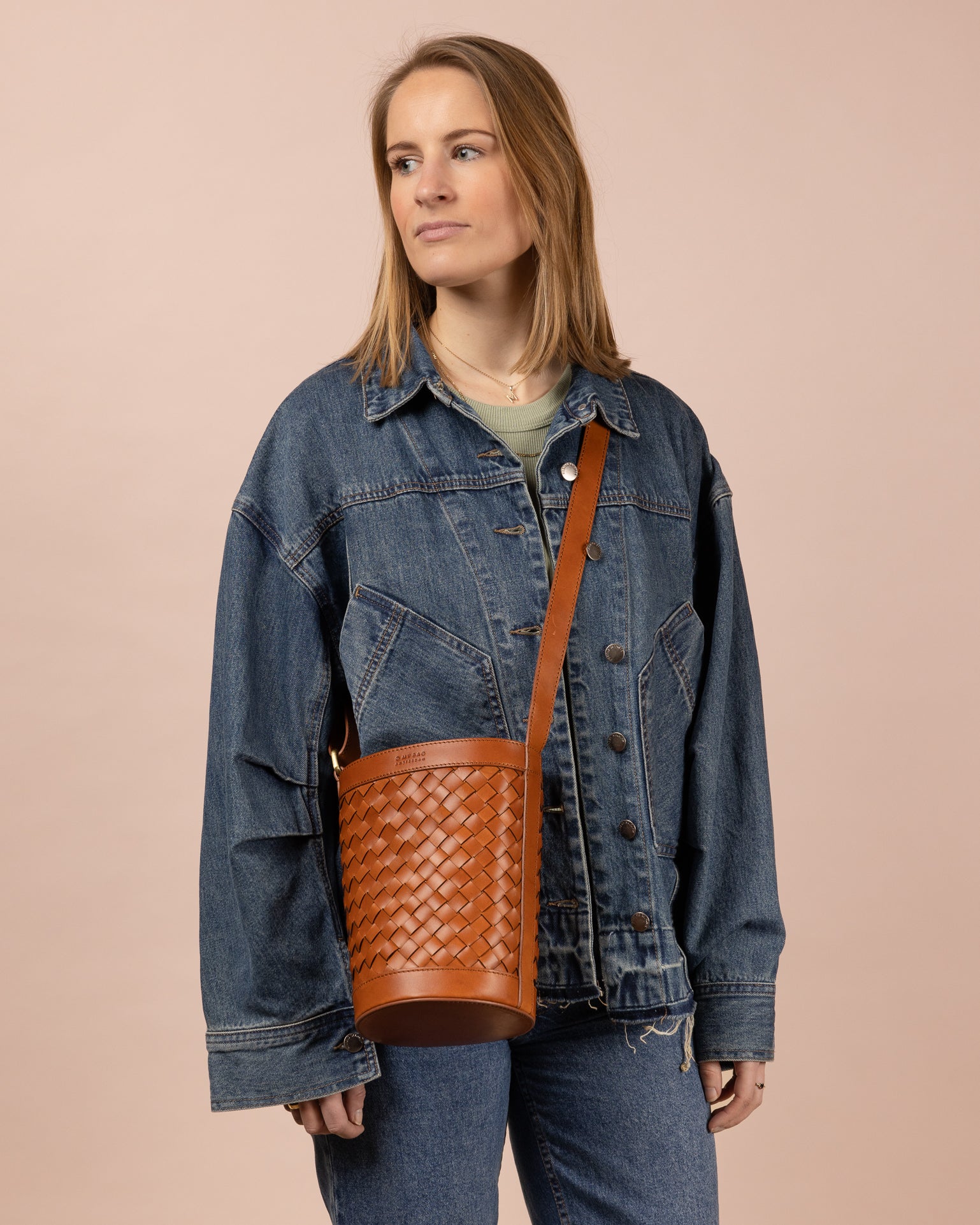 Image of model holding circular woven bucket bag in cognac leather. Cross-body shot.