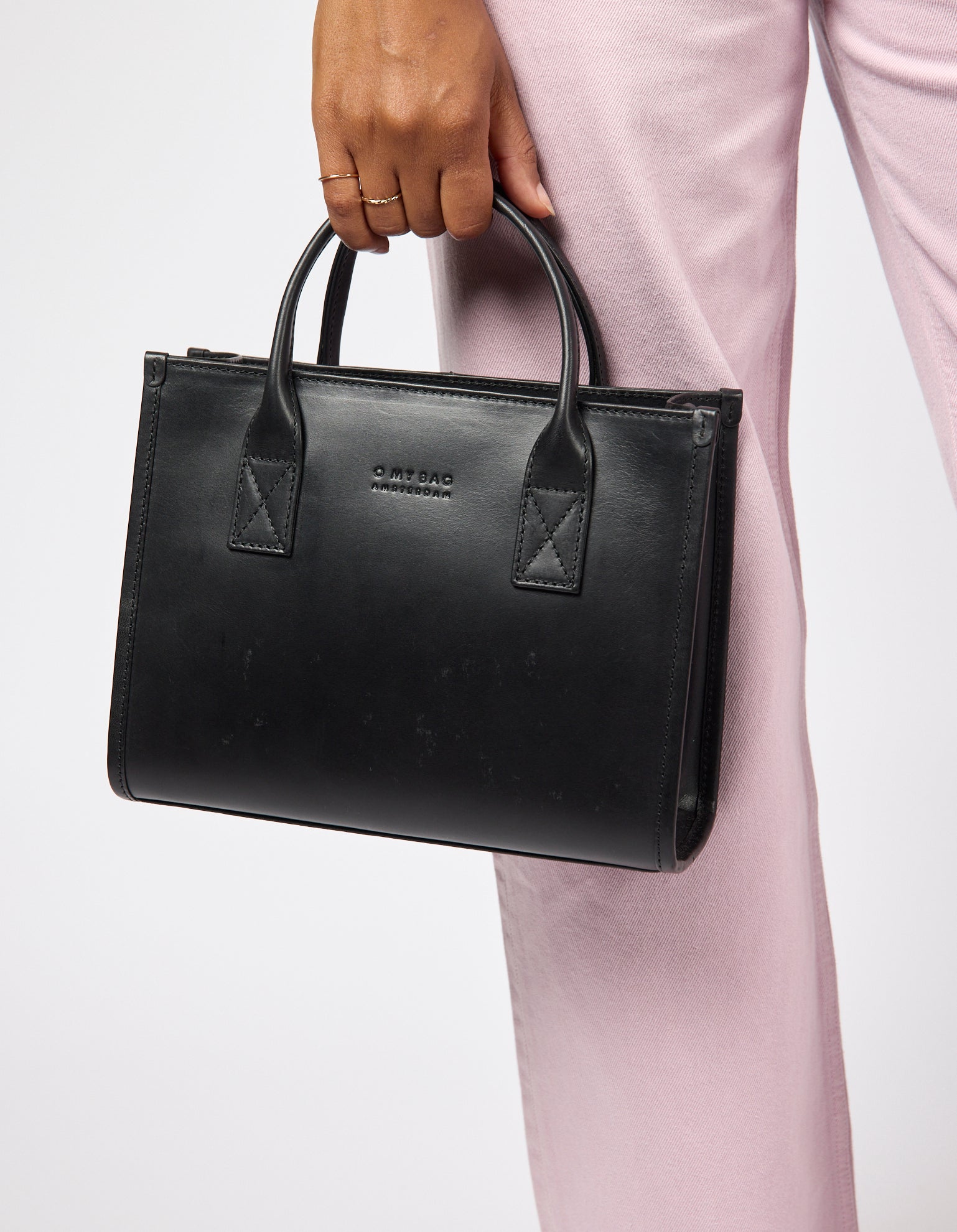 Black handbag held by a person wearing pink pants on a white background