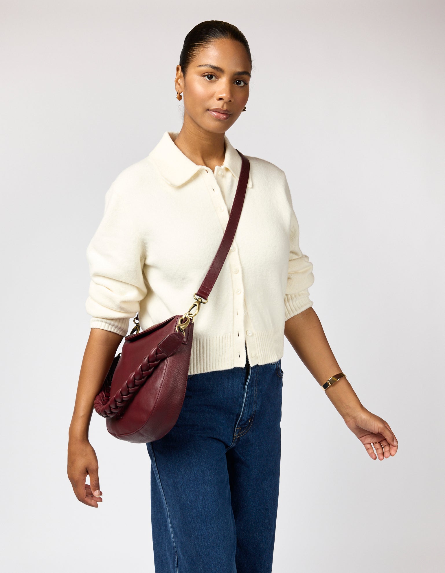Woman wearing a white sweater and blue jeans, holding a maroon handbag against a plain background