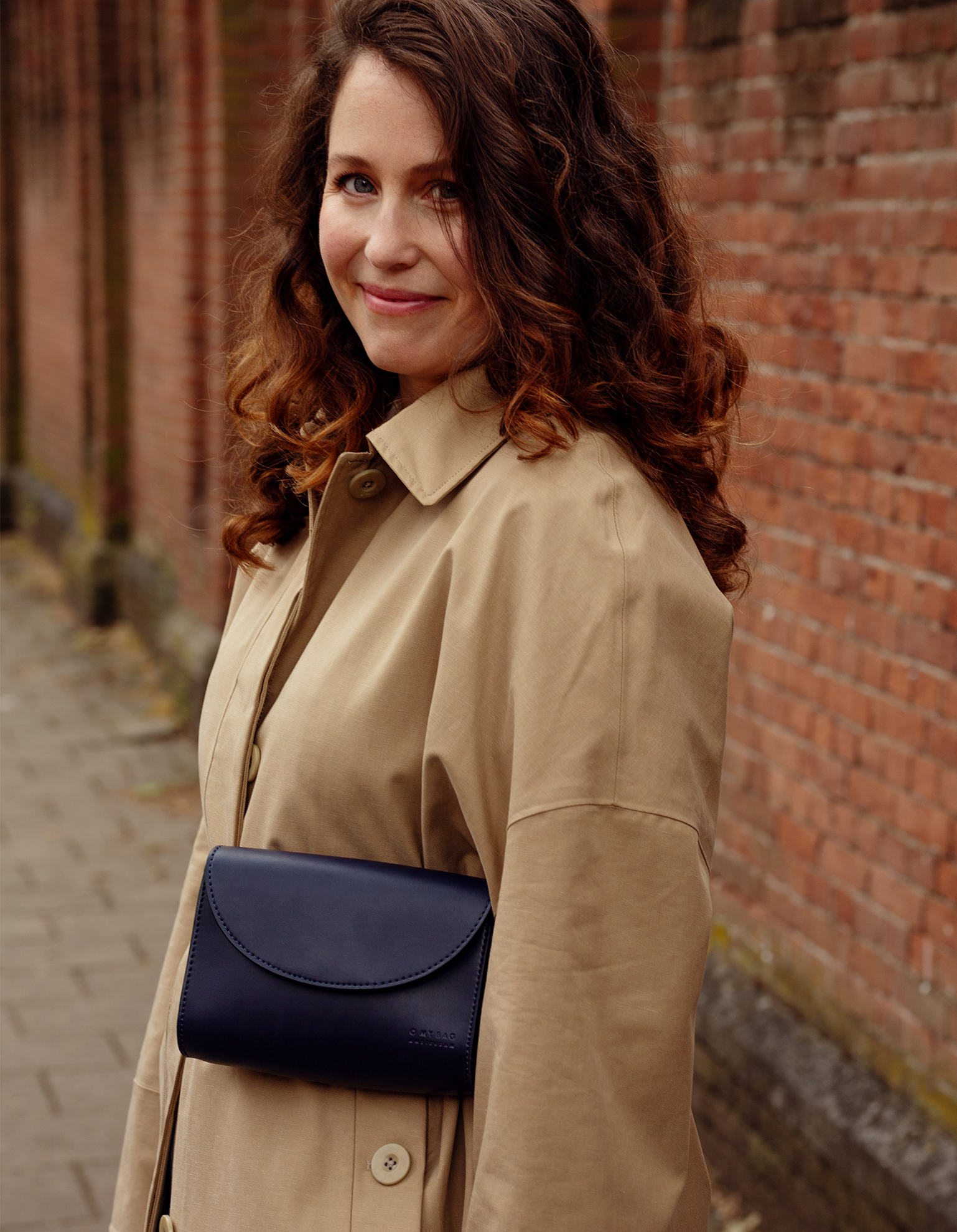 Campaign image of woman with brown hair wearing navy belt bag over a trench coat