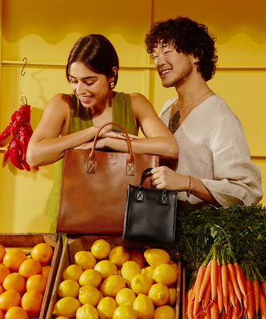 Two models holding mini and maxi versions of a tote bag in front of a market stall