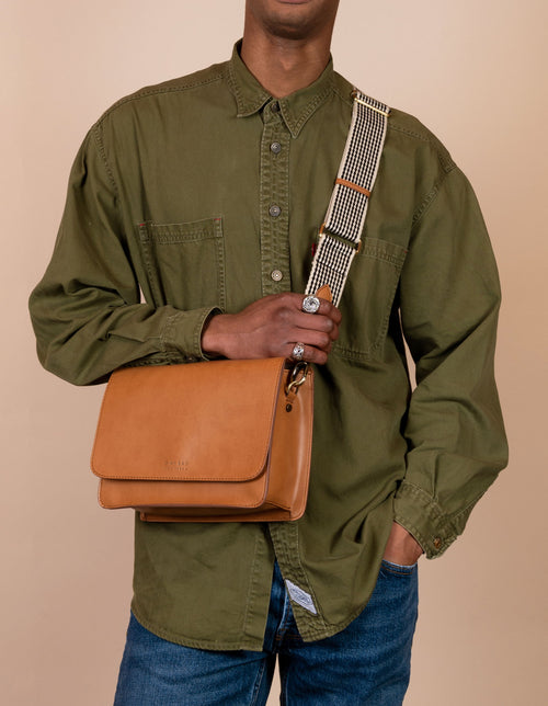 Male model with Audrey in cognac apple leather and checked webbing strap.