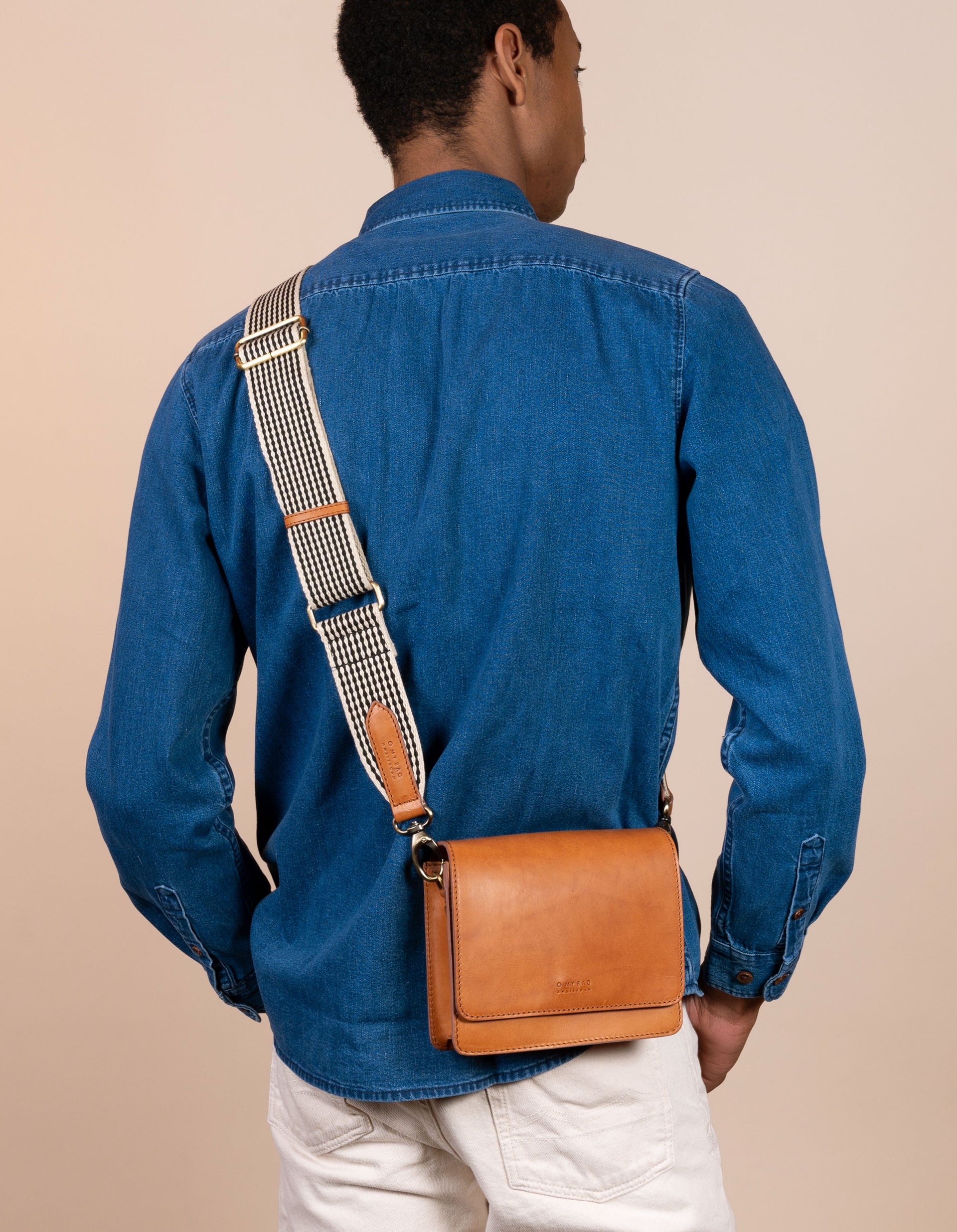 Audrey Mini Cognac leather bag. Square shape with an adjustable webbing strap. Male Model image. Back view